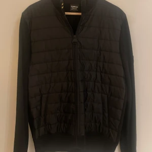 Svart quiltad cardigan från Barbour - Snygg svart quiltad cardigan från Barbour International med dragkedja framtill. Jackan har ribbade detaljer vid kragen och ett märke på ärmen. Perfekt för en stilren look. Storlek M tts, 2 innerfickor respektive ytterfickor. Nypris runt 2200kr.