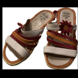 Färgglada sandaler - Säljer ett par Scholl sandaler i färgerna vit, röd och orange. Sandalerna har en dekorativ blomma på remmen och flera färgglada remmar för en unik look. Perfekta för sommarens äventyr! 