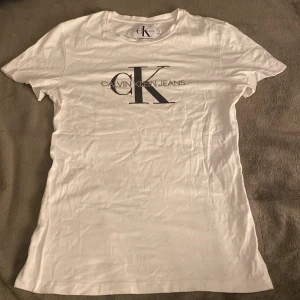 Vit t-shirt från Calvin Klein Jeans - Säljer en stilren vit t-shirt från Calvin Klein Jeans med det ikoniska CK-logotypen tryckt på framsidan. T-shirten är kortärmad och tillverkad i mjuk bomull, nästan aldrig använt den så tröjan är i ett bra skick 