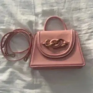 Snygg rosa handväska med en elegant kedjedetalj på framsidan. Väskan har ett handtag och en avtagbar axelrem för flexibilitet. Perfekt för att ge en färgklick till din outfit!