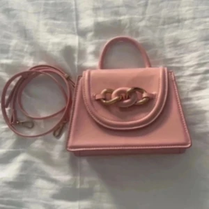 Rosa handväska med kedjedetalj - Snygg rosa handväska med en elegant kedjedetalj på framsidan. Väskan har ett handtag och en avtagbar axelrem för flexibilitet. Perfekt för att ge en färgklick till din outfit!
