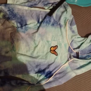 Tie-dye hoodie med fjärilsdetalj - Säljer en färgglad tie-dye hoodie med en broderad fjäril på bröstet. Hoodien har en justerbar dragsko i huvan och långa ärmar. Perfekt för en avslappnad stil.