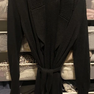 Svart stickad kappa från Sisley - Elegant svart stickad kappa från Sisley med knytband i midjan. Den har långa ärmar och en stilren design som passar perfekt för kyligare dagar.