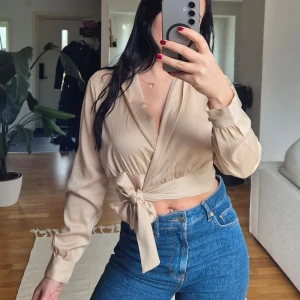 Beige omlottopp från Bubbleroom - Säljer en elegant beige omlottopp från Bubbleroom i samarbete med Sandra Willer. Toppen har långa ärmar och en knytning i midjan för en smickrande passform. Perfekt för en stilren look.