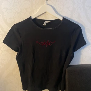 Svart ribbad topp från H&M - Snygg svart ribbad topp från H&M med korta ärmar och ett coolt rött broderi framtill. Perfekt för en stilren look. Har gjort broderi själv så de är one of its kind:)