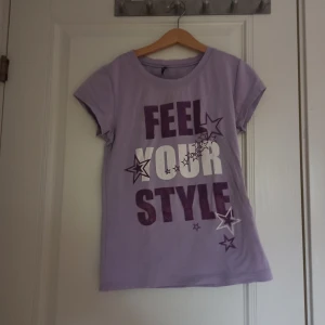 Lila t-shirt med text och stjärnor - Snygg lila t-shirt med texten 'FEEL YOUR STYLE' i vitt och lila. Dekorerad med stjärnor i olika storlekar. Perfekt för en y2k stil och vintage stil. 