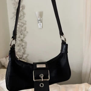 Svart axelväska från Dolce & Gabbana - Snygg svart axelväska från Dolce & Gabbana med stilren design. Väskan har ett silver spänne, grå inuti med en innerficka. Köpte den från Plick och köparen kunde inte garantera äkthet, därav priset! Väskan är däremot i ett otroligt fint skick!