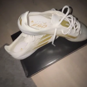 Vita och guldiga fotbollsskor från Adidas - Snygga Adidas F50 fotbollsskor i vitt med guldiga detaljer. Skorna har snörning och är perfekta för fotbollsplanen. De har en modern design med de klassiska tre ränderna i guld på sidan. Helt nya, perfekt skick. Elite.