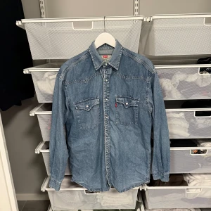 Blå jeansskjorta från Levi's - Snygg blå jeansskjorta från Levi's med klassisk krage och två bröstfickor med knappar. Perfekt för en avslappnad stil. Skjortan har långa ärmar och en rak passform. Storlek M