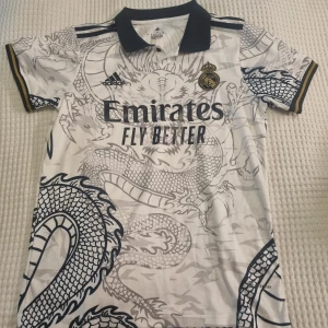 Vit fotbollströja från Adidas - Vit fotbollströja från Adidas med drakmönster. Tröjan har korta ärmar och en mörblåbkrage. Real Madrid-logga och sponsortryck på framsidan.