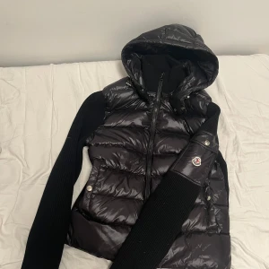 Svart cardigan från Moncler - Snygg svart cardigan från Moncler med huva och dragkedja. Jackan har långa ärmar i ett ribbat material och en glansig finish på kroppen. Perfekt för kyligare dagar. Storlek L med passar m bäst.