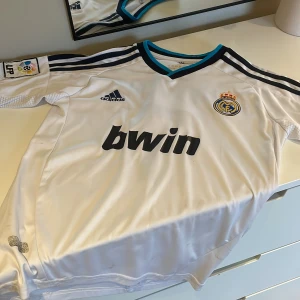 Vit Real Madrid fotbollströja från Adidas - Säljer en vit retro Real Madrid fotbollströja med svart och blå detaljer. Tröjan har korta ärmar och är prydd med 'Ronaldo 7' på ryggen. Den har även LFP-loggan på ärmen och är tillverkad i ClimaCool-material för optimal komfort. Den är från säsongen 2012/2013. Den är använd några gånger och lite text har suddats ut men är ändå i bra skick. Den är i storlek 12. 
