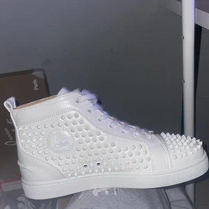 Vita sneakers med nitar från Christian Louboutin - Snygga vita sneakers från Christian Louboutin med nitar över hela ovandelen och den ikoniska röda sulan. Skorna har snörning och en högre modell som ger en cool look.