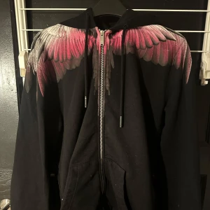 Svart hoodie med vingmönster från Marcelo Burlon - Snygg svart hoodie från Marcelo Burlon med ett unikt vingmönster i rosa och grått över axlarna. Tröjan har dragkedja framtill och en bekväm huva. Perfekt för en cool och avslappnad stil.
