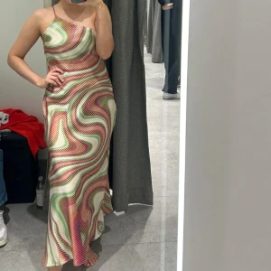 Färgglad långklänning med mönster - Snygg långklänning med ett färgglatt, vågigt mönster i grönt, rött och beige. Klänningen har en smal axelbandslös design som ger en elegant look. Perfekt för sommarens festligheter!