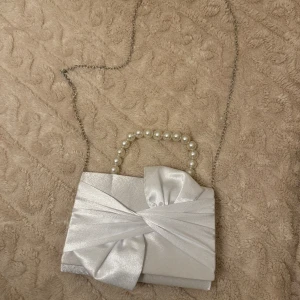 Vit väska - Elegant vit väska i satin med en unik rosettdetalj på framsidan. Väskan har en kedja för axelrem och ett handtag dekorerat med pärlor, perfekt för speciella tillfällen.