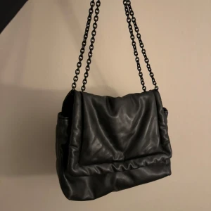 Svart axelväska med kedja - Snygg svart axelväska i skinnimitation med kedjeaxelremmar. Väskan har en mjuk och slouchy design, perfekt för en trendig look. Den är rymlig och passar till många olika stilar.