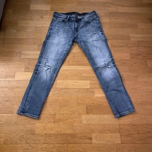 Blå jeans med slitningar - Säljer ett par blå jeans med coola slitningar och en klassisk femficksdesign. De har en knapp och dragkedja framtill. Perfekta för en avslappnad stil. MIDJA 33 LÄNGD 32
