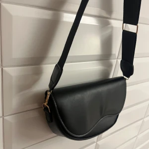 Svart axelväska med justerbar rem från SHEIN eller Temu  - Snygg svart axelväska i syntetmaterial med justerbar rem och magnetlås. Perfekt för att bära dina nödvändigheter med stil. Enkel och elegant design som passar till många tillfällen. Aldrig använd. Säljer för 50 kronor 