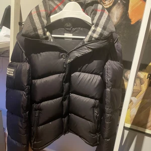 Burberry jacka - Säljer en snygg svart pufferjacka från Burberry med klassiskt rutigt foder i huvan. Jackan har dragkedja och knappar framtill samt en broderad logga på ärmen. Perfekt för kyliga dagar, och även inför våren då man kan ta av armarna och göra till väst. Kvitto finns!                                                              Hör av er vid fler frågor💛🤗