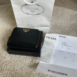 Svart plånbok från Prada - Snygg och elegant svart plånbok från Prada i skinn med dragkedja. Perfekt för att hålla ordning på kort och kontanter. Kommer med originalförpackning och kvitto.