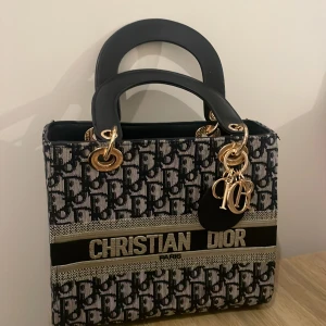 Mönstrad handväska  - Snygg handväska  med ett ikoniskt mönster i svart och beige. Väskan har guldfärgade detaljer och ett elegant handtag. Perfekt för att ge din outfit en lyxig touch.