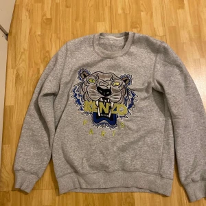 Grå sweatshirt från Kenzo med tigertryck - Säljer en grå sweatshirt från Kenzo med ett stort tigertryck på framsidan. Tröjan har långa ärmar och rund halsringning. Perfekt för en avslappnad stil med en touch av lyx.