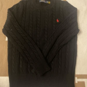 Svart stickad tröja från Polo Ralph Lauren - Säljer en klassisk svart stickad tröja från Polo Ralph Lauren med kabelstickat mönster och det ikoniska röda logotypbroderiet på bröstet. Tröjan är för liten för mig, kvitto finns och även taggen på tröjan sitter kvar 
