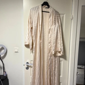 Beige kimono med randigt mönster - Elegant beige kimono med diskret randigt mönster. Klänningen har trekvartsärmar och en luftig passform, perfekt för en stilren look. Tillverkad i ett lätt och bekvämt material som ger en fin drapering.