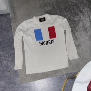 Vit tröja från Morris - Säljer en vit tröja från Morris med tryck i blått och rött på framsidan. Tröjan har långa ärmar och en rund halsringning. Perfekt för en stilren look.