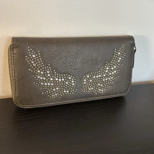 Grå plånbok med vingdetaljer - Snygg grå plånbok med glittriga vingdetaljer på framsidan. Plånboken har flera fack och en dragkedja runtom för säker förvaring. Perfekt för att hålla ordning på kort och kontanter.