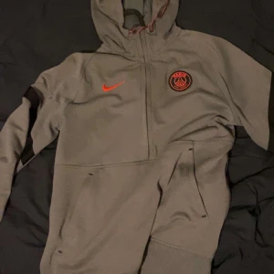 Grå hoodie från Nike med PSG-logga - Snygg grå hoodie från Nike med Paris Saint-Germain-logga på bröstet. Hoodien har en orange Nike-swoosh och en praktisk ficka framtill. Perfekt för kyliga dagar. Vid snabb affär pris säkning 