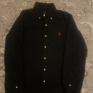 Svart skjorta från Ralph Lauren - Snygg svart skjorta från Ralph Lauren med klassisk krage och knappar framtill. Den har en röd broderad logotyp på bröstet och är långärmad. Perfekt för en stilren look, nypris 1400, pris är inte hugget i sten vid snabb affär! Köpt på ROOM 1006 butik. Strl S. Inga defekter! Mer lik en M/L i storleken pga längd