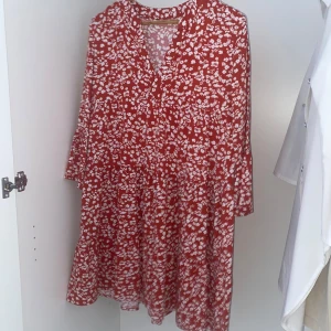 Röd blommig klänning - Charmig röd klänning med vita blommor och v-ringad hals. Den har långa ärmar med en lätt volangdetalj vid ärmsluten. Perfekt för en somrig look! 