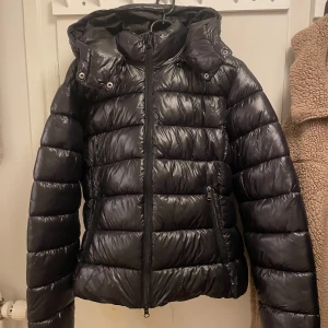 Svart pufferjacka från 157 - Säljer en svart pufferjacka från 157 med huva och dragkedja. Jackan har en glansig yta och är perfekt för kyligare väder.                                     Den är i storlek L men skulle säga att den sitter som en M, jag har endast använt den 1 gång och sen dess har den bara hängt i garderoben därför vill jag nu sälja den