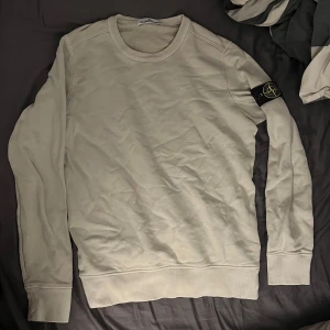 Beige tröja från Stone Island - Säljer en stilren beige tröja från Stone Island med rund hals och långärmad design. Tröjan har en ikonisk logga på ärmen och är perfekt för en avslappnad look. Passar bra till både jeans och chinos. Tröjan är 10/10 skick och säljer den då för att den inte blivit använd på länge. Tröjan är äkte och har även en qr kod där man kan checka att den är autentisk. Pris kan diskuteras vid snabb affär.