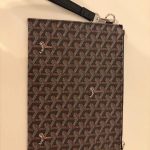 Brun mönstrad kuvertväska - Snygg brun kuvertväska med ett unikt geometriskt mönster. Väskan har en praktisk dragkedja och en avtagbar handledsrem. Perfekt för att bära det viktigaste med stil.
