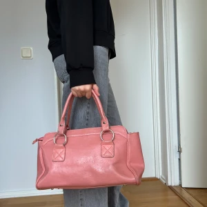 Rosa handväska med dragkedja - Säljer en snygg rosa handväska med dragkedja och handtag. Väskan har stilrena detaljer med ringar i metall och den har två innerfack. Väskan är i en praktisk storlek för vardagsbruk. Perfekt för att bära med sig det viktigaste med stil. 