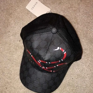 Svart keps från Gucci med ormmönster - Snygg svart keps från Gucci med ett iögonfallande rött och vitt ormmönster. Kepsen har ett diskret mönster i tyget och justerbar passform. Perfekt för att ge din outfit en lyxig touch.
