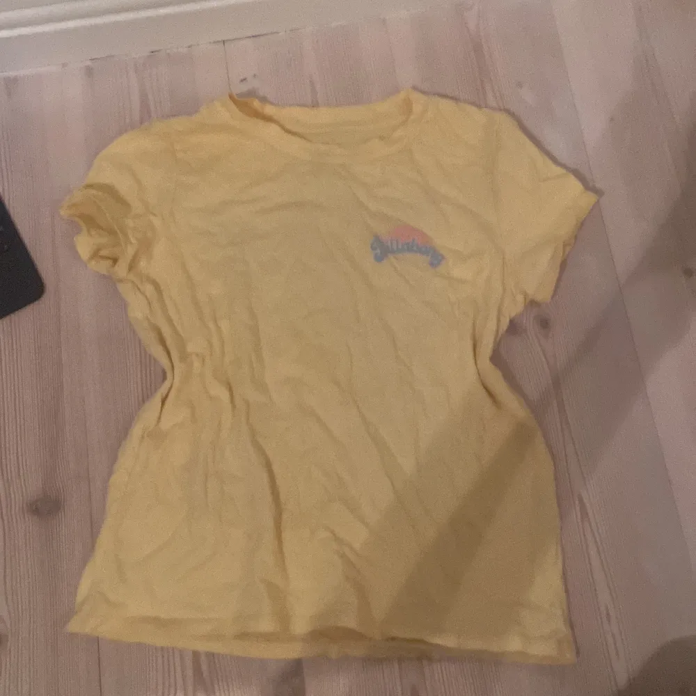 Säljer en gul t-shirt från Billabong med ett tryck av en solnedgång och palmer på baksidan. T-shirten har korta ärmar och en avslappnad passform. Perfekt för sommardagar!. T-paidat.
