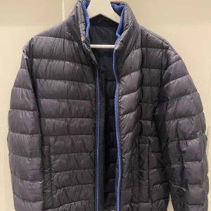 Mörkblå dunjacka från Hugo Boss - Snygg mörkblå dunjacka från Hugo Boss med blå detaljer.Fyllning av 80% ankdun och 20% fjäder. Sitter som en Medium