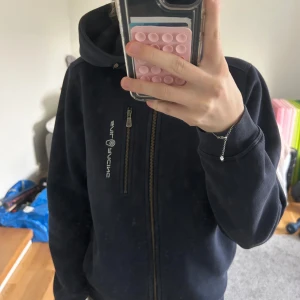 Mörkblå hoodie från Sail Racing - Säljer en mörkblå hoodie från Sail Racing med dragkedja och huva. Jackan har en bröstficka med dragkedja och logotyp på bröstet och ärmen. Storlek 160. Nypris ca 1000kr 