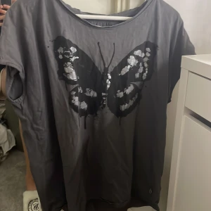 Grå t-shirt med fjärilstryck - Snygg grå t-shirt med ett stort fjärilstryck i svart och silver på framsidan. T-shirten har en avslappnad passform och korta ärmar, perfekt för en casual look. 🦋