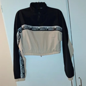 Svart och beige vindjacka från Adidas - Säljer en snygg svart och beige vindjacka från Adidas med dragkedja och elastisk nederkant. Jackan har långa ärmar och ett coolt band med Adidas-logga längs ärmarna och bröstet. Perfekt för en sportig look!