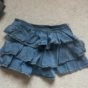 Blå denim volangkjol från Blanco - Charmig blå volangkjol från Blanco i jeansmaterial. Kjolen har flera lager av volanger och en dragkedja vid sidan för enkel påklädning. Coolt att ha kedjan framåt också! Midje: 36cm rakt över, Längd 32cm