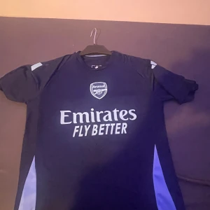 Blå Arsenal fotbollströja - Snygg blå Arsenal fotbollströja med korta ärmar och vit logga på bröstet. Perfekt för fans som vill visa sitt stöd. Tröjan har en modern design med ljusblå detaljer på sidorna.