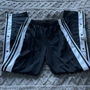 Byxor - Snygga svarta byxor från Adidas med vita ränder och tryckknappar längs sidorna. Byxorna har Adidas logga på vänster ben. Midja med dragsko för att kunna justera passformen. Använd fåtal gånger🤗