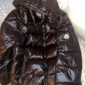 Svart pufferjacka från Moncler - Snygg svart pufferjacka från Moncler med glansig finish och huva. Jackan har en dragkedja framtill och en ficka med Moncler-logga på ärmen. Perfekt för kyliga dagar.