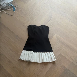 Svart vit klänning  - Elegant svart axelbandslös klänning med en kontrasterande vit plisserad kjol. Perfekt för en stilren look med en touch av klassisk elegans.
