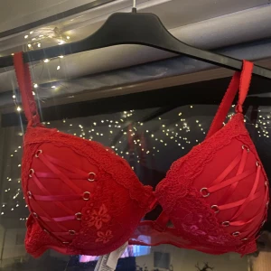 Röd spets-bh med snörning - Säljer en snygg röd bh med spetsdetaljer och snörning framtill. Den har justerbara axelband och en vacker blommig spetsdesign. Perfekt för en festlig kväll eller när du vill känna dig extra fin. Med avtagbara axelband.